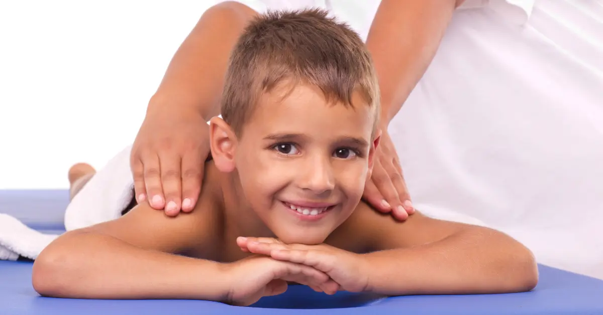 Smiling boy lying on a massage table while a therapist's hands rest on his shoulders