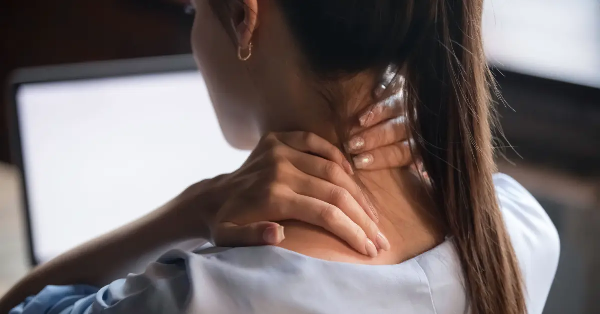 Woman at a laptop pressing her fingers into a tight spot at the back of her neck