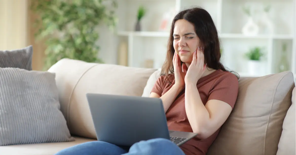 Woman on a couch with a laptop, wincing and pressing both hands to her jaw